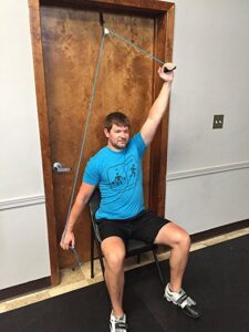 A person performing a shoulder‑pulley exercise using an over‑the‑door hook system, demonstrating assisted mobility and range‑of‑motion work with scratch‑resistant door protection.