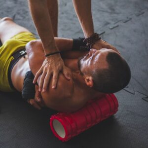 A person lying on a gym floor using a foam roller under the upper back while assisted by a trainer, demonstrating physiotherapy‑style muscle release and recovery techniques.
