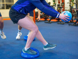 Athlete balancing on one leg on a wobble cushion while reaching for a med ball during agility training